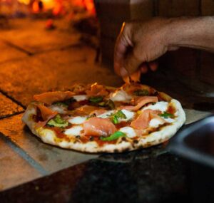 Cook placing ingredients on pizza in the mouth of the wood fired oven - pizza in Aldinga