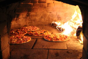 5 pizzas bake on one side of the wood-fired oven, logs ablaze on the other side.